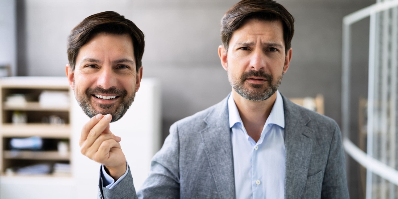 A white man with short dark hair, moustache and stubble and wearing a grey jacket, looks morose while holding up a life sized cut out photograph of his face in which he is smiling. The background is an office with a cabinet.